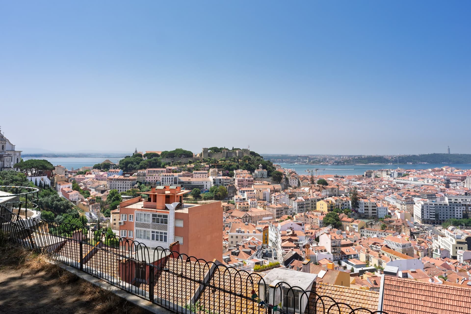 A wide view across Lisbon's pale rooftops from a hilltop terrace, with the castle walls on the left.