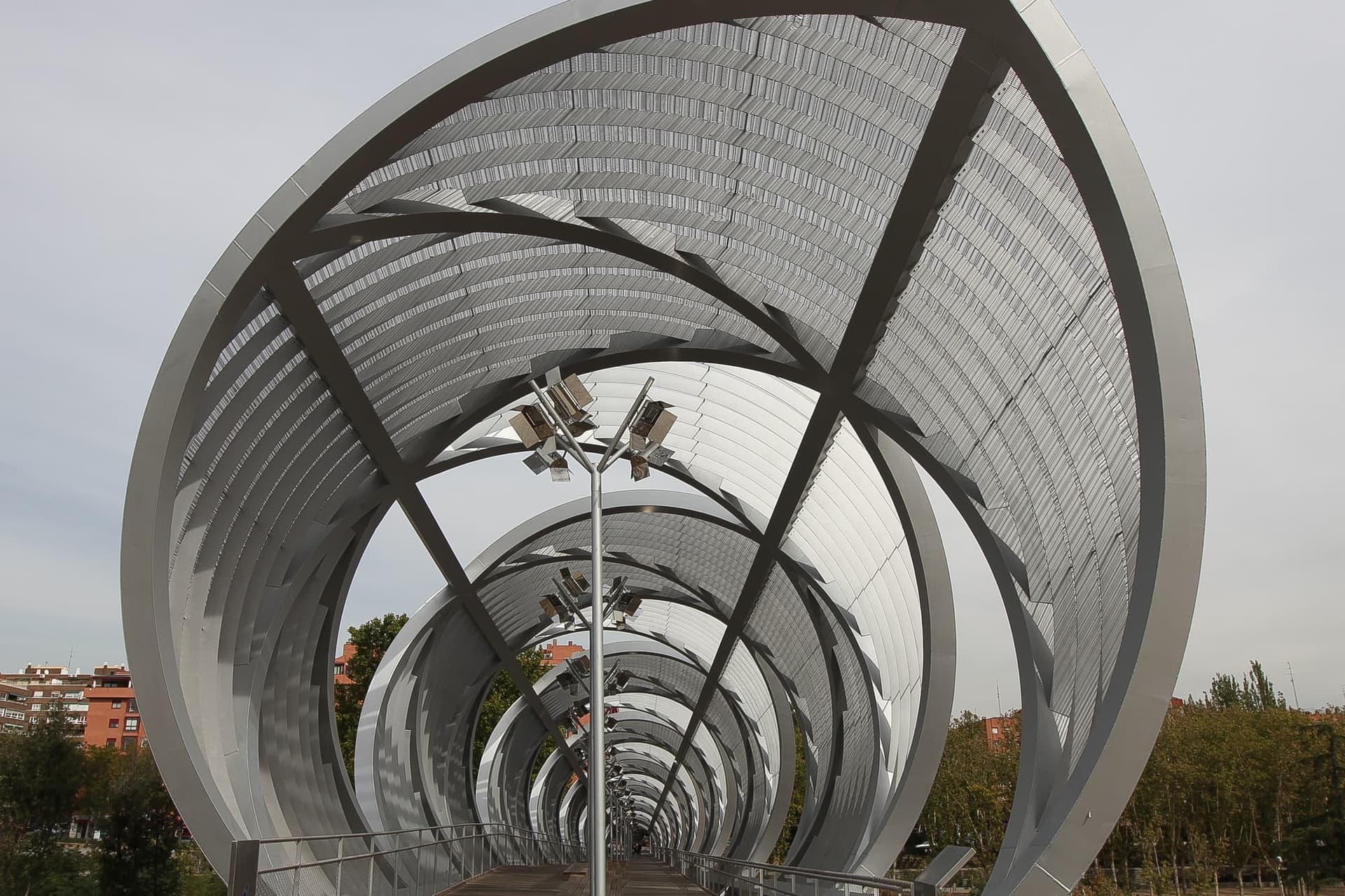 The spiral steel footbridge over the Manzanares river in Madrid Río park, with the city behind it.